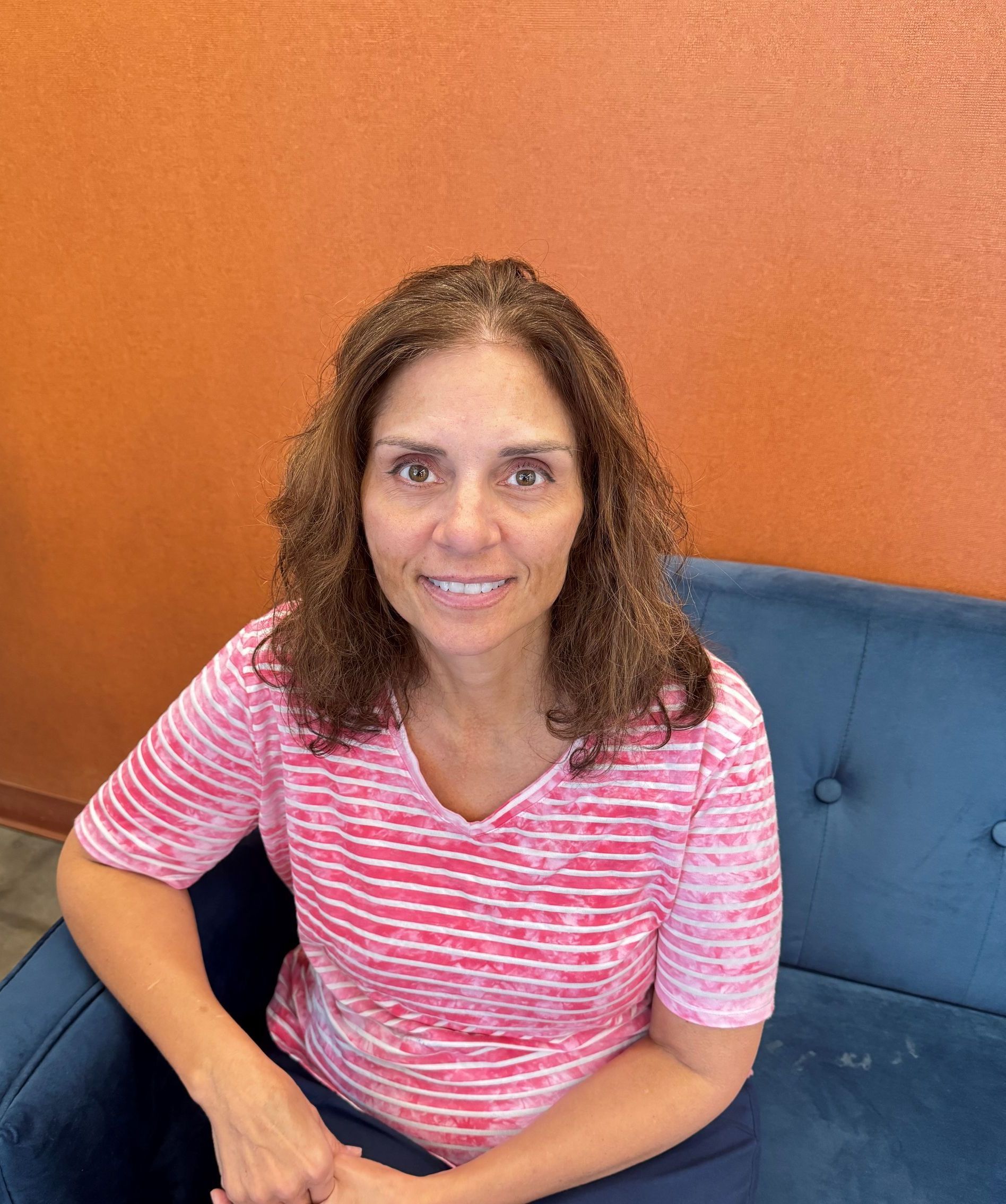 Sharon Alexander with curly brown hair in a pink striped shirt seated on a blue couch against an orange wall.