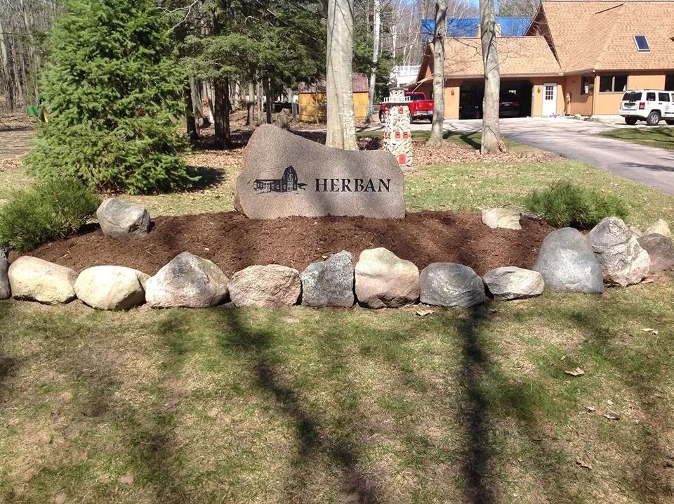 Sign for Herban, a stone monument with carved text and silhouette, surrounded by landscaping, trees, and a house.