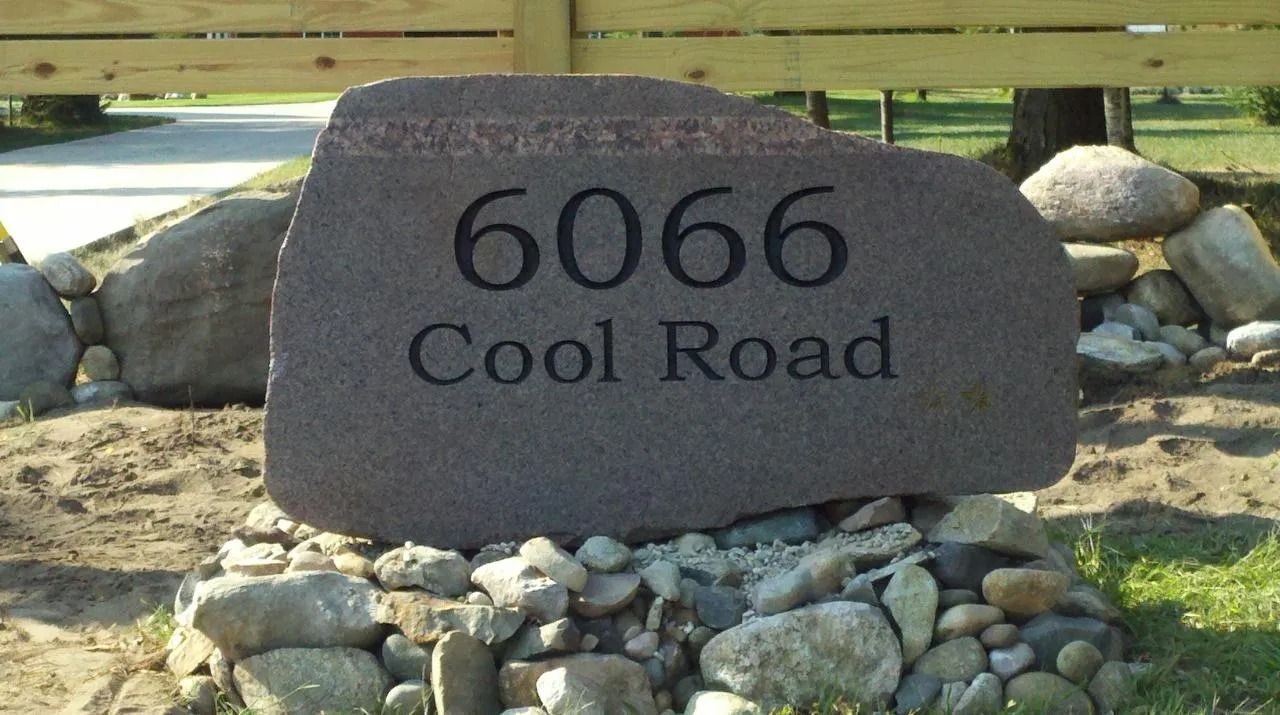 Address marker: 6066 Cool Road, engraved on a large, brown stone set atop smaller rocks, with wooden fence in background.