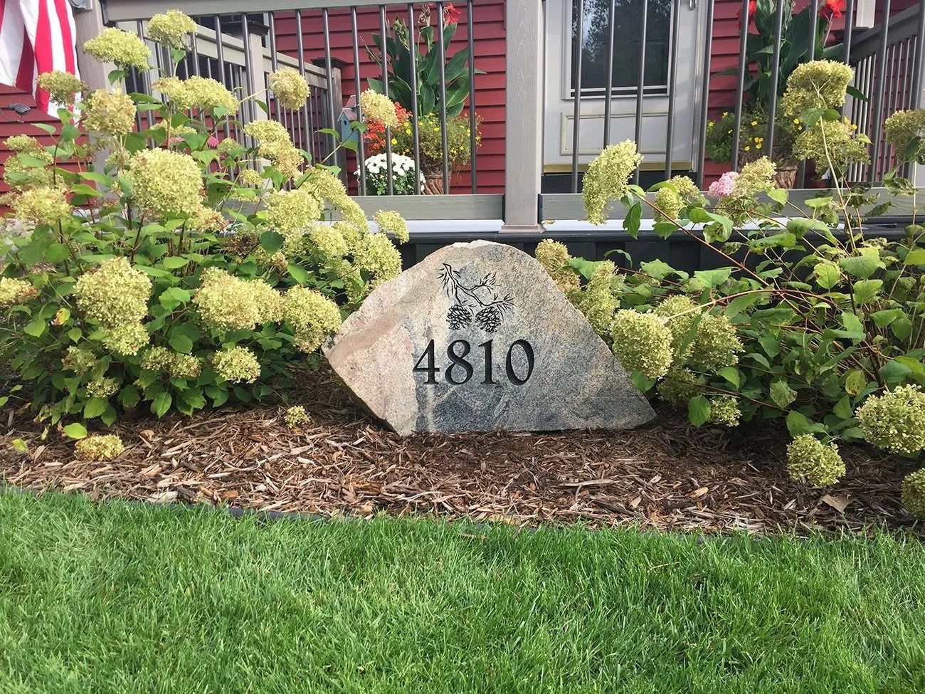 Rock with house number 4810, surrounded by green shrubs and mulch, in front of a red building with a porch.