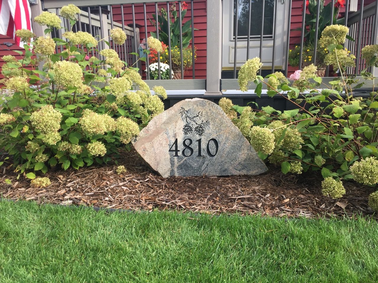 A large rock with the house number 4810 engraved sits in front of a red house, surrounded by plants.