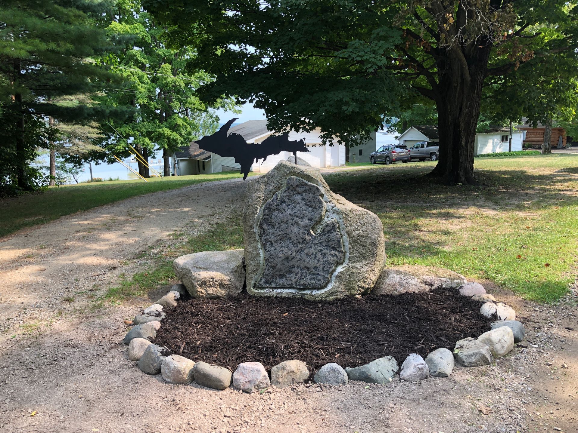 Large rock with Michigan's Upper Peninsula silhouette. Stone circle border. Gravel path and trees in background.
