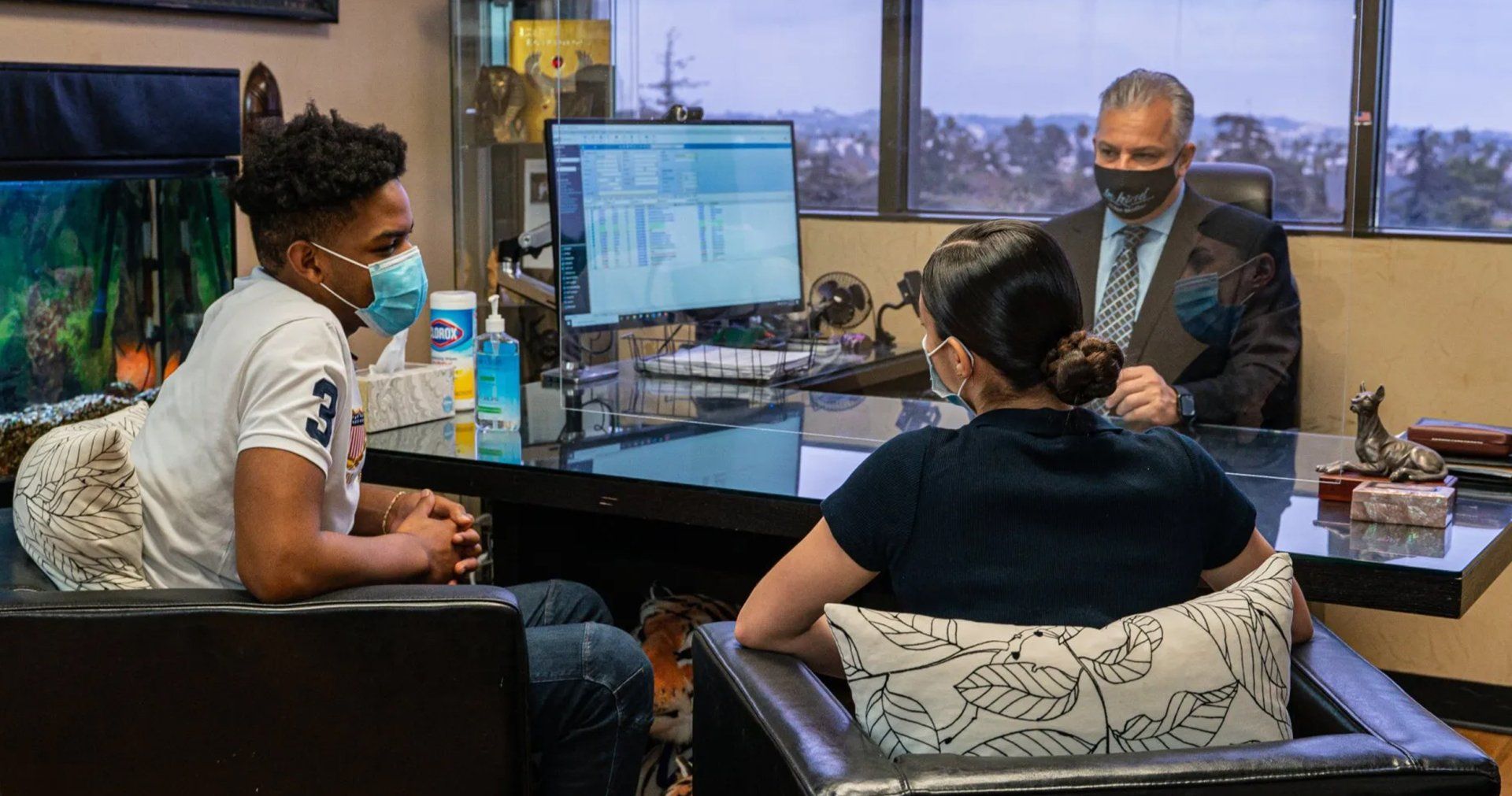 Three people wearing masks in an office. A man in a suit sits behind a desk. Two others sit in chairs.