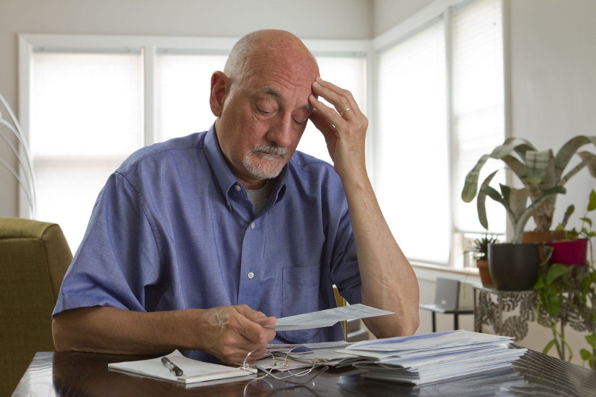 Man with hand on forehead, looking at bills, appearing stressed. Desk with papers, pen, windows.