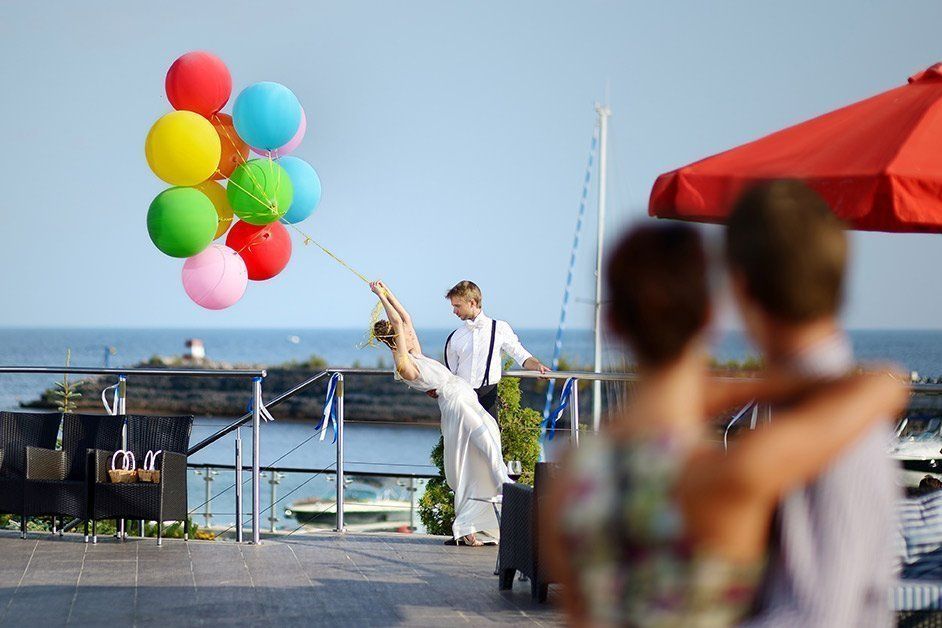Woman held aloft by colorful balloons, man watching on outdoor patio by the sea.