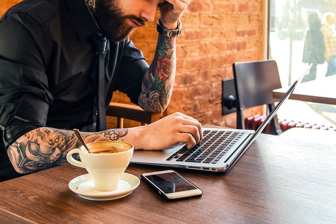 Man with tattoos, stressed, typing on laptop in cafe. Coffee and phone on table.