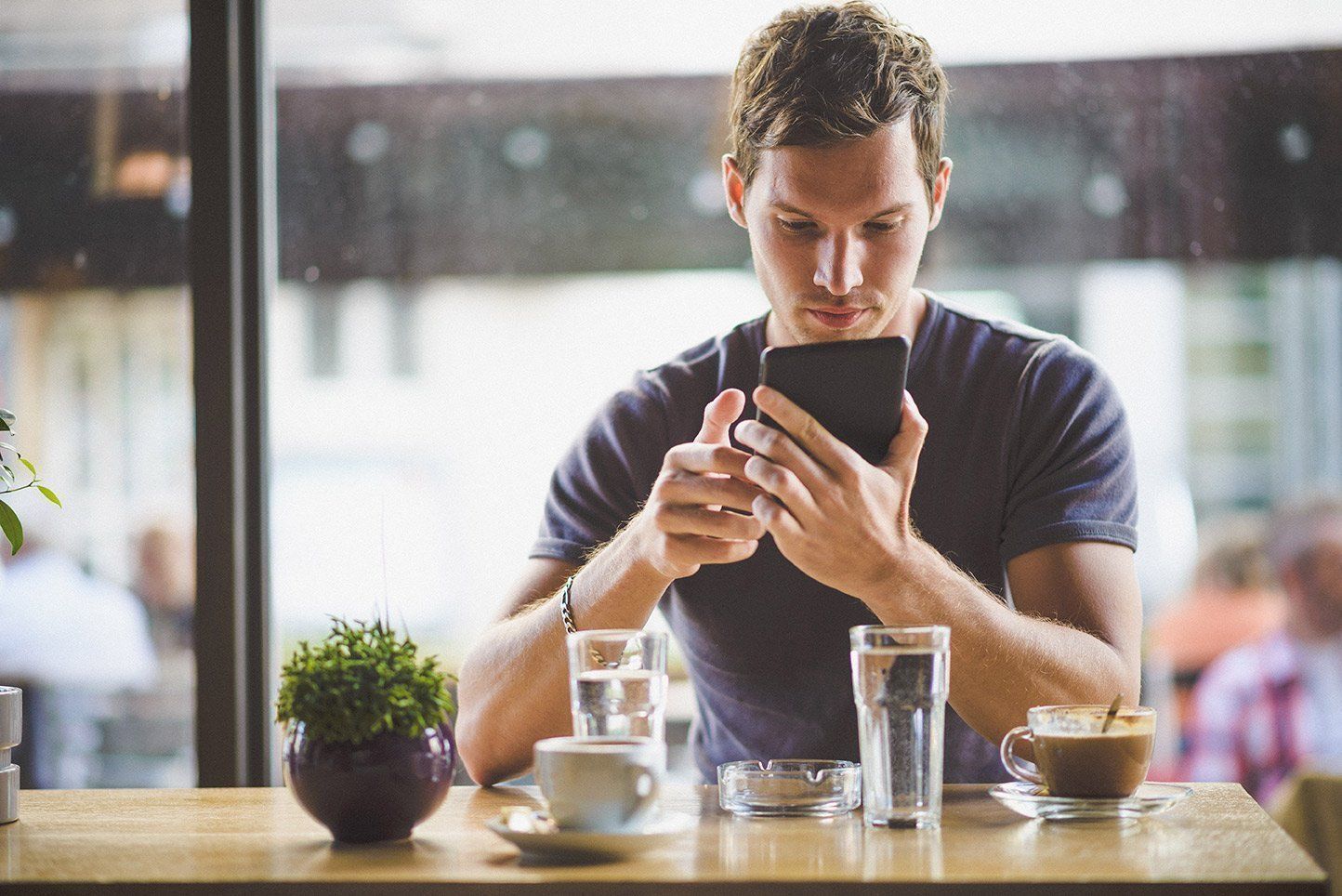 Man using a phone at a cafe table. He is looking down intently. Coffee cup and water glass on the table.