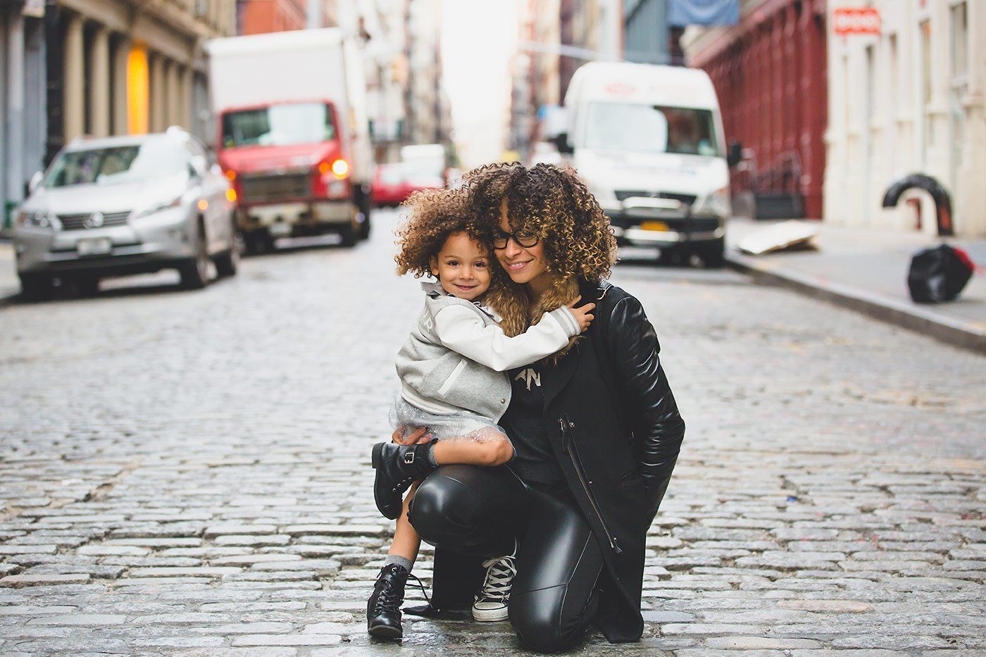 Woman and child embrace on a cobblestone street with vehicles in the background.