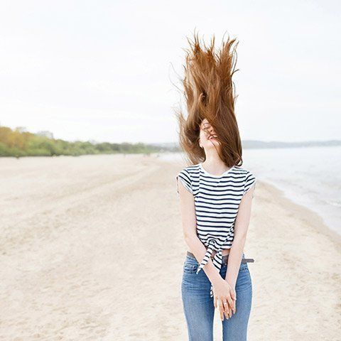 Woman flips long brown hair on beach, wearing striped shirt and jeans.