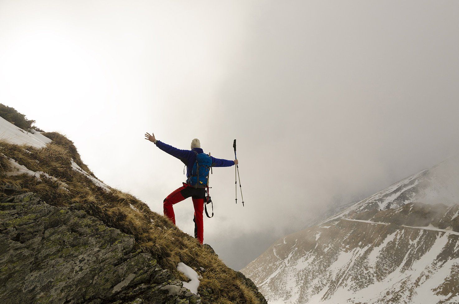 Mountaineer on rocky peak with arms raised, holding trekking poles. Snowy mountain backdrop, cloudy sky.