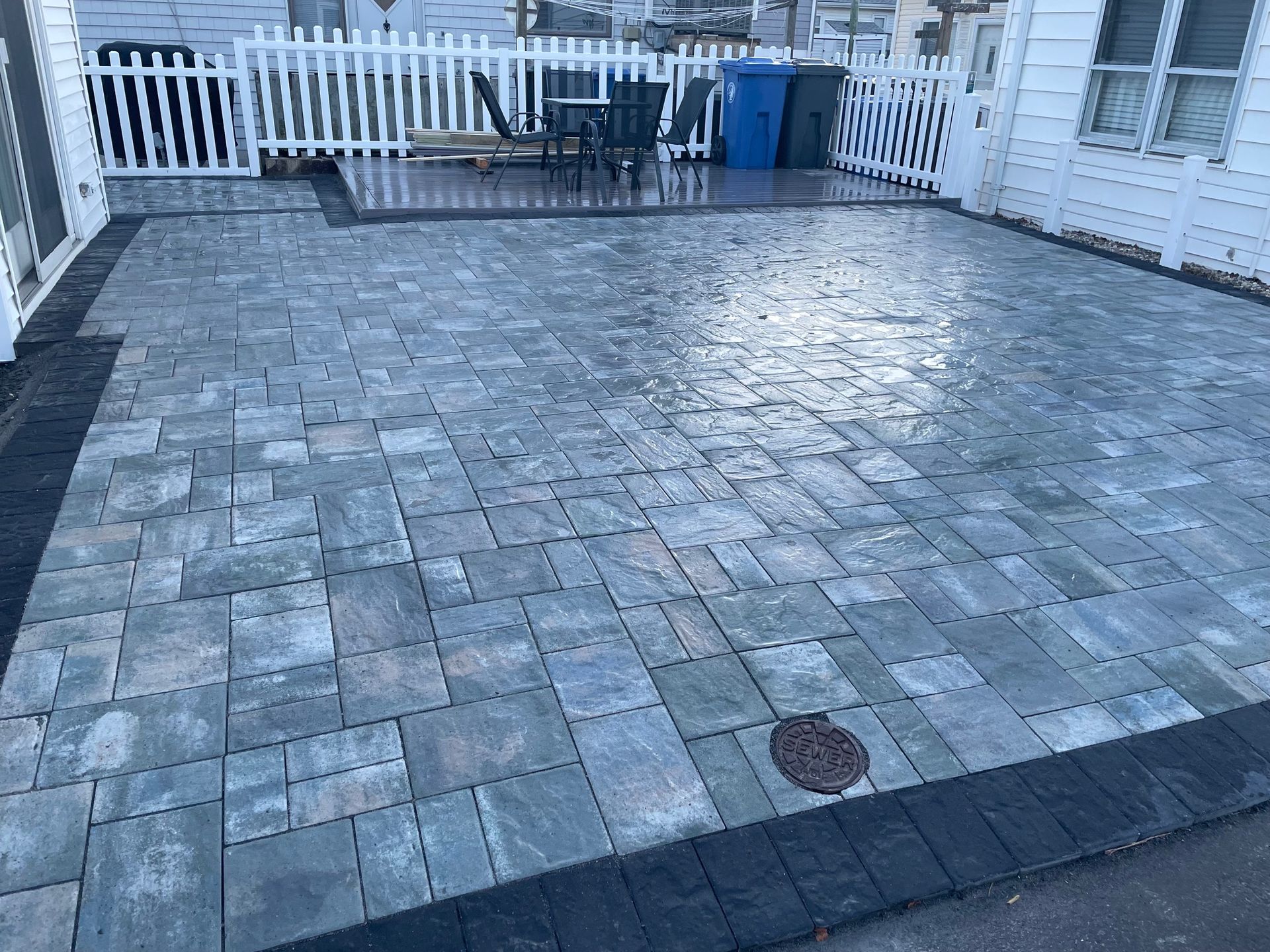 Paver patio with dark border next to a white house and fence.