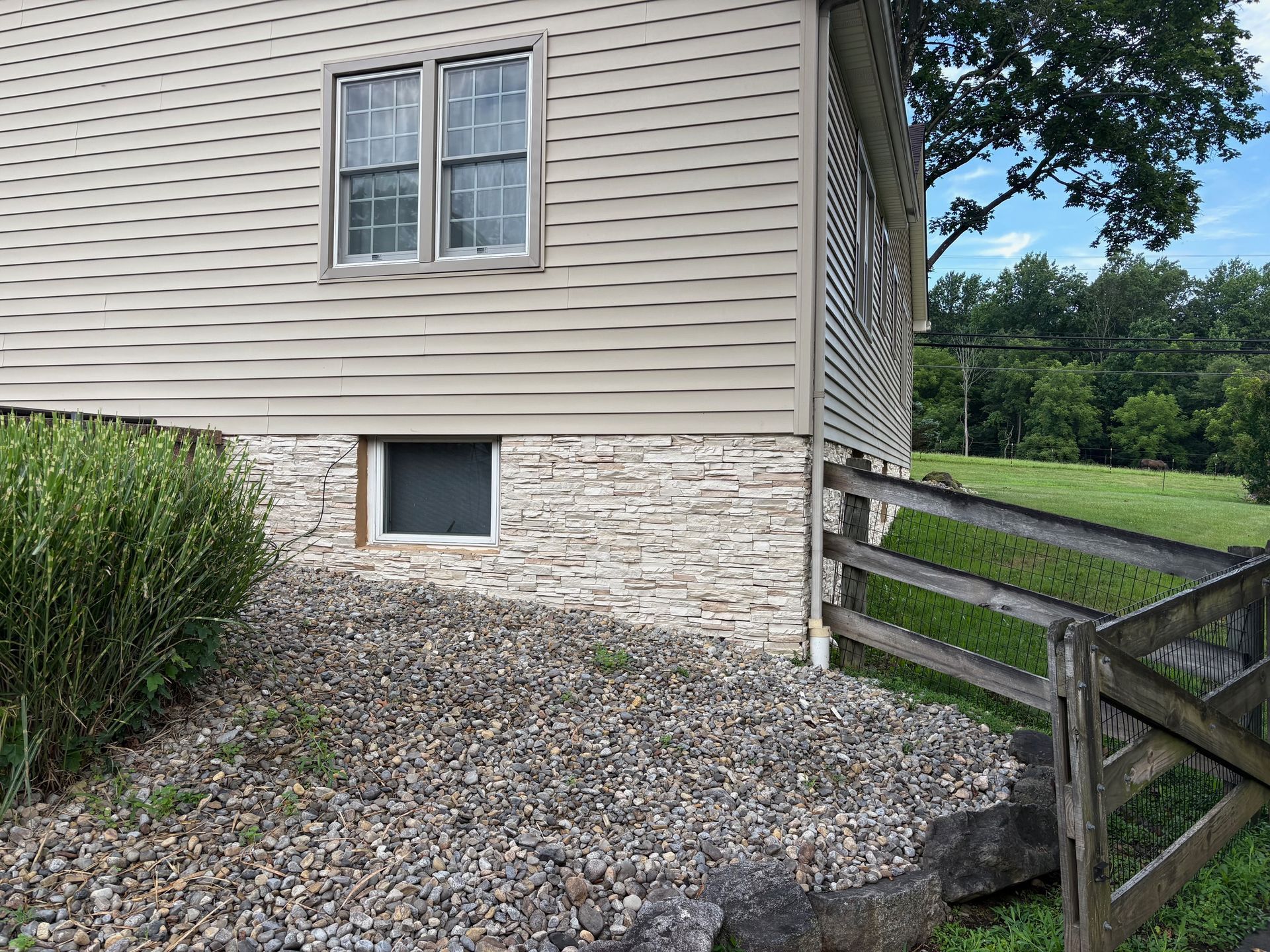 Stone and siding house exterior, window, and small fence with a grassy yard and trees.