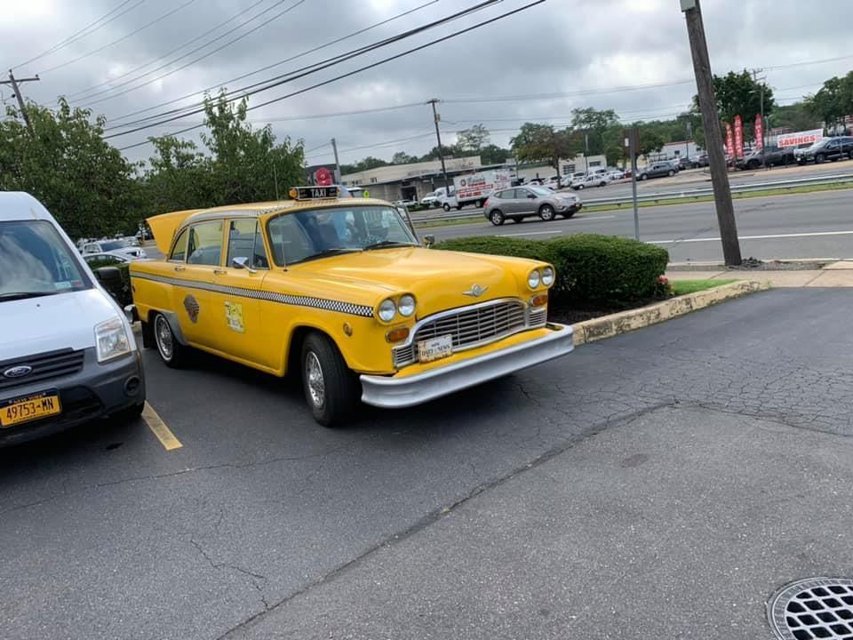 Yellow vintage car parked in a lot beside a white van, with a street and cloudy sky in the background