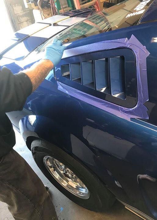 Person masking a blue car panel around a vent opening in a workshop.