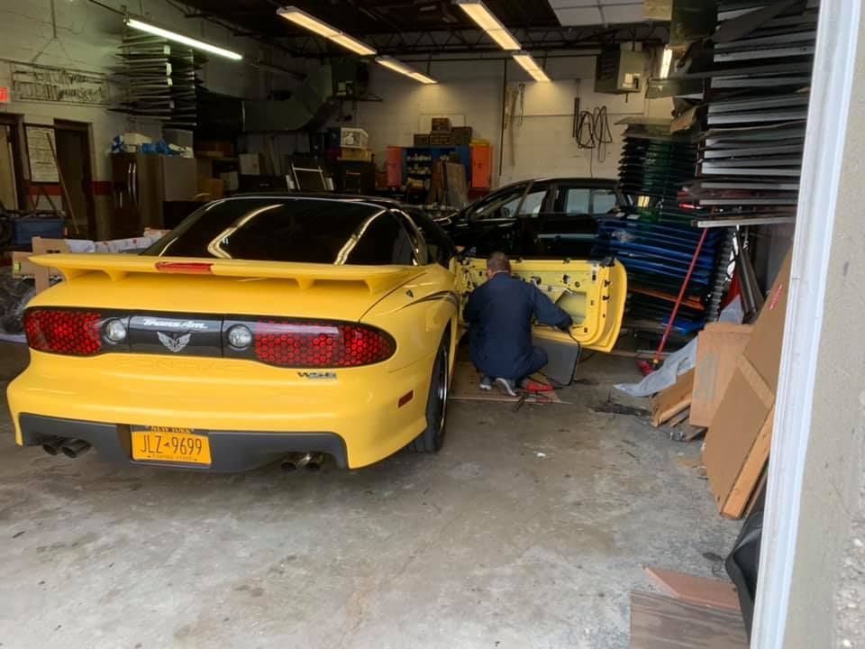 Yellow sports car in a garage with a mechanic working beside it and stacked parts nearby