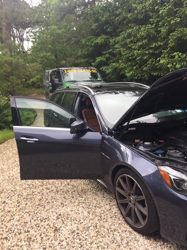 Dark blue car with open hood and driver door on a gravel drive, with another vehicle behind it in trees.