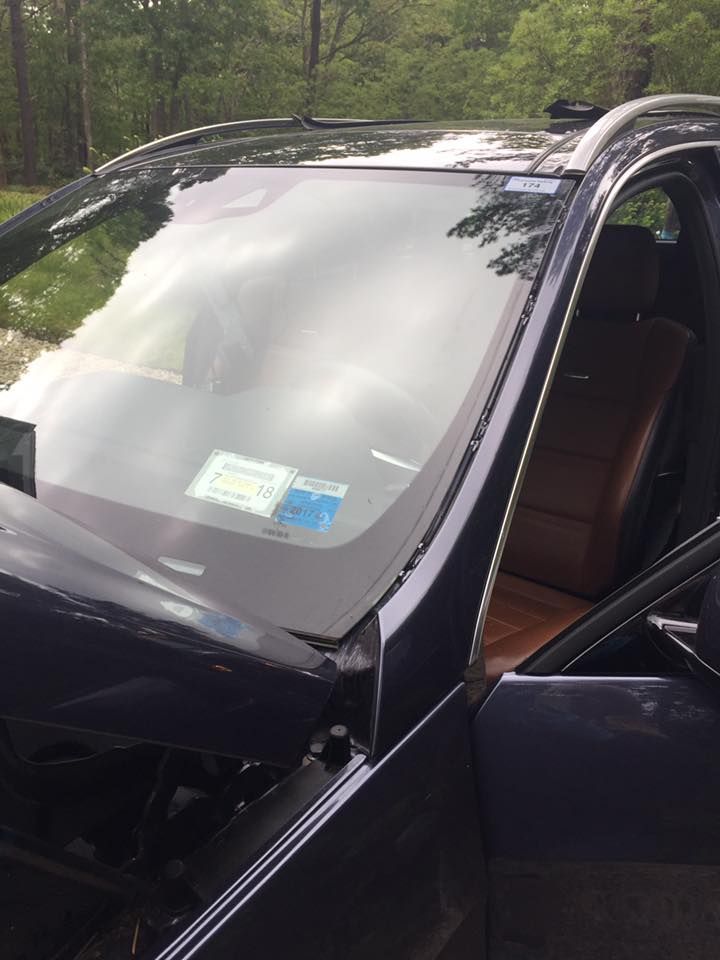 Black car with open driver door and cracked front windshield in a wooded area
