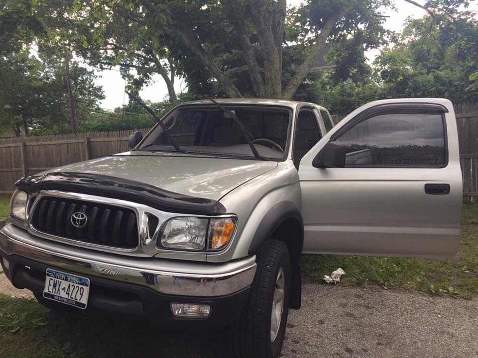 Silver Toyota pickup with open passenger door parked on a gravel driveway beside trees and a wooden fence