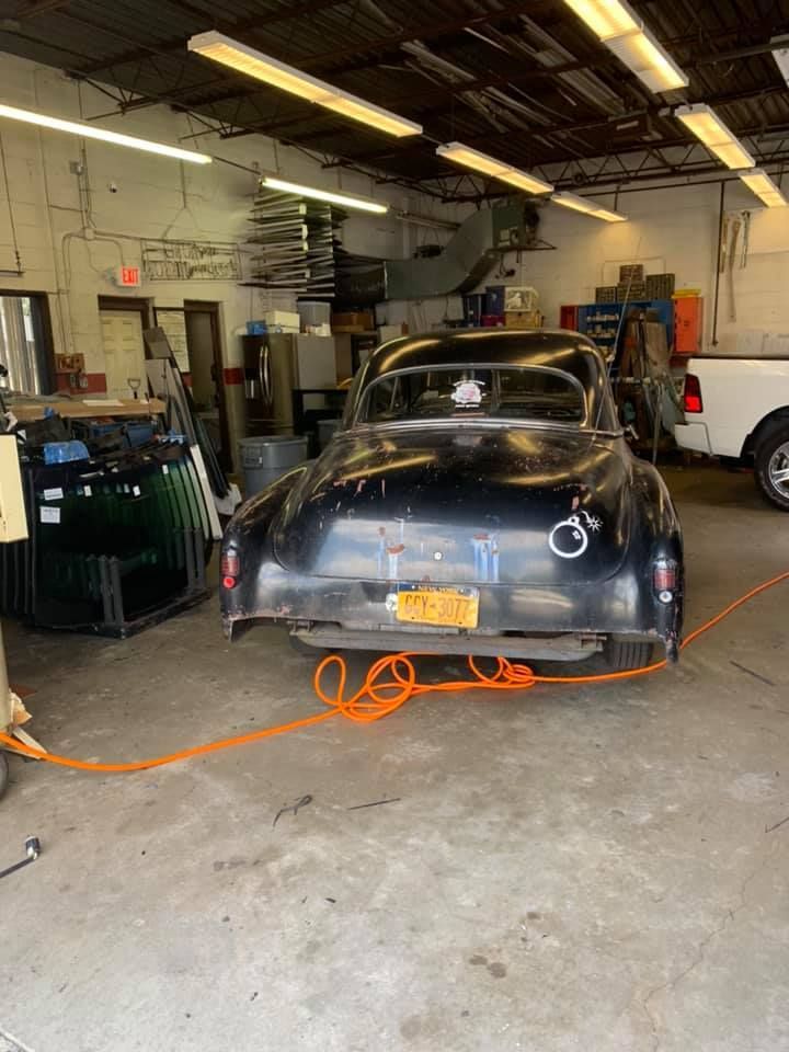 Black vintage car in a cluttered auto shop, with an orange extension cord on the floor.