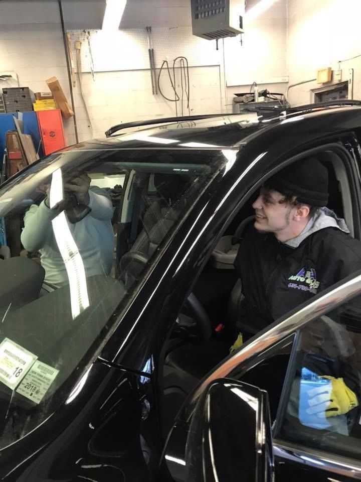 Person in a black car at an auto shop, leaning toward the open door while a mechanic works nearby