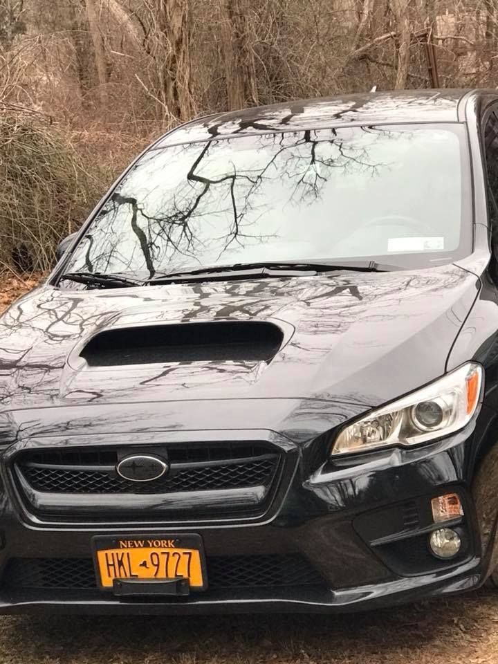 Black sedan with a hood scoop parked on a dirt roadside, front view with tree reflections on the windshield.