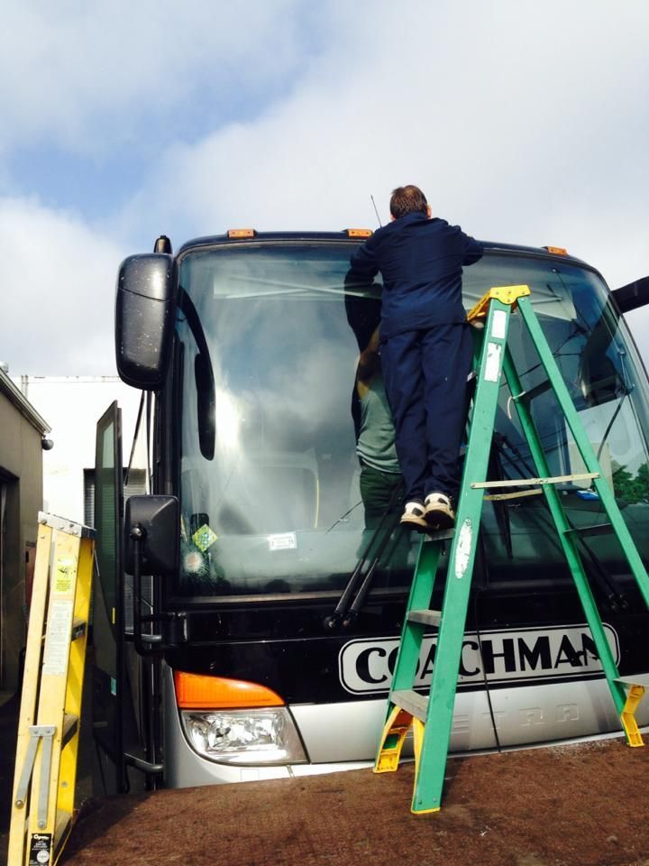 Person on green ladder repairing windshield of a Coachman bus outdoors