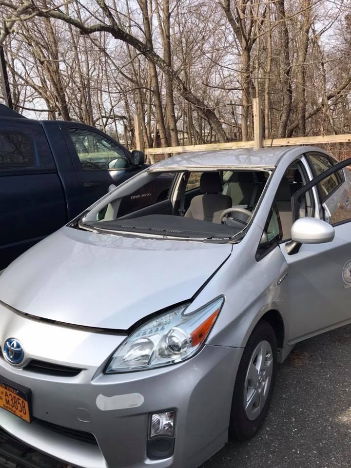 Silver car with shattered windshield and missing side windows parked beside a dark SUV in a wooded lot