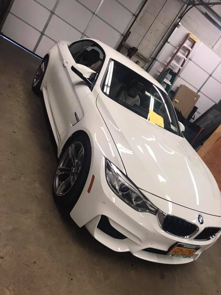 White BMW coupe in a garage, angled front view with black wheels and yellow plate.