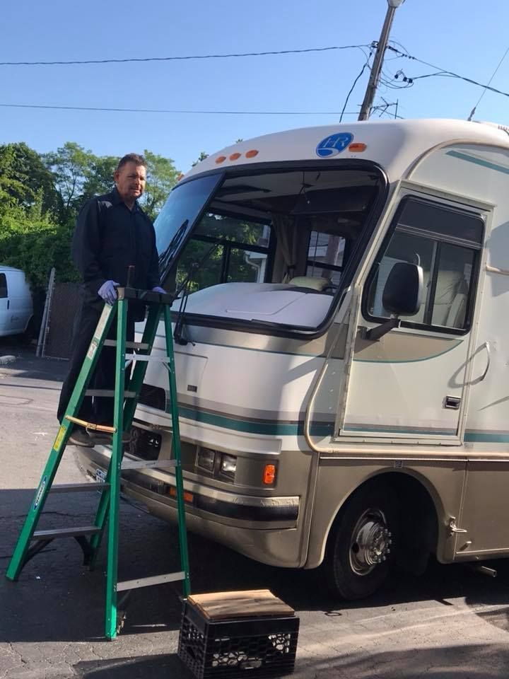 Man standing on a green ladder beside a white RV, working near the windshield in a sunny driveway