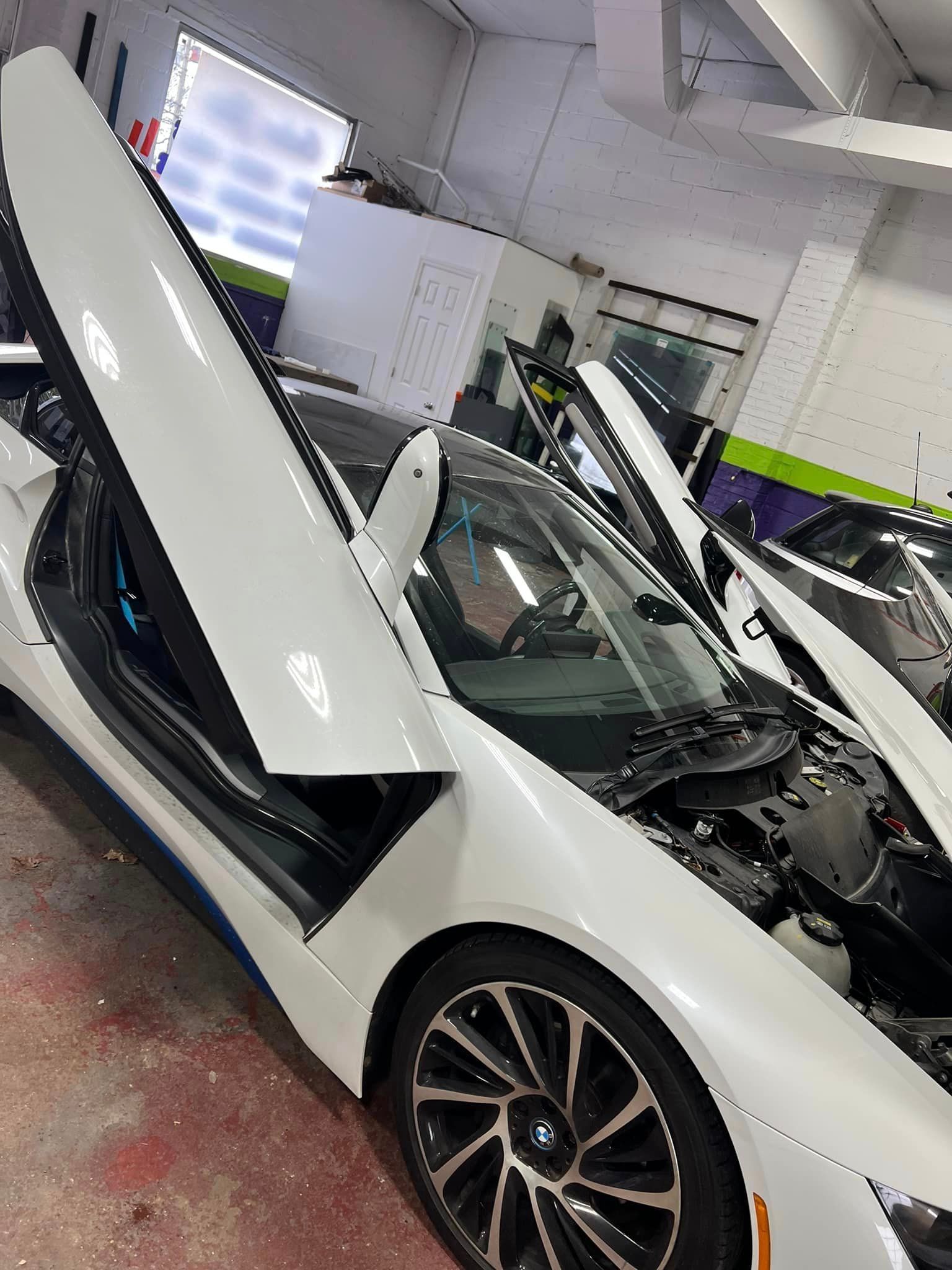 White sports car in a workshop with both gullwing-style doors open and hood raised.