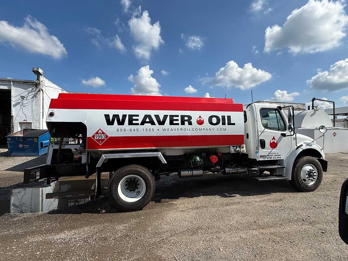 A Weaver Oil fuel truck with red and white paint, parked on gravel against a blue sky.