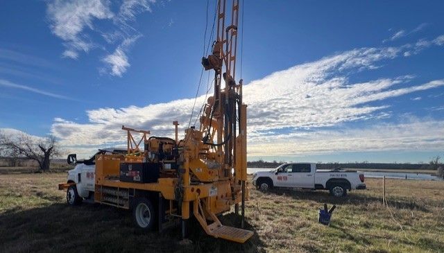 Excavator driving a pile with a work truck parked on a construction site.