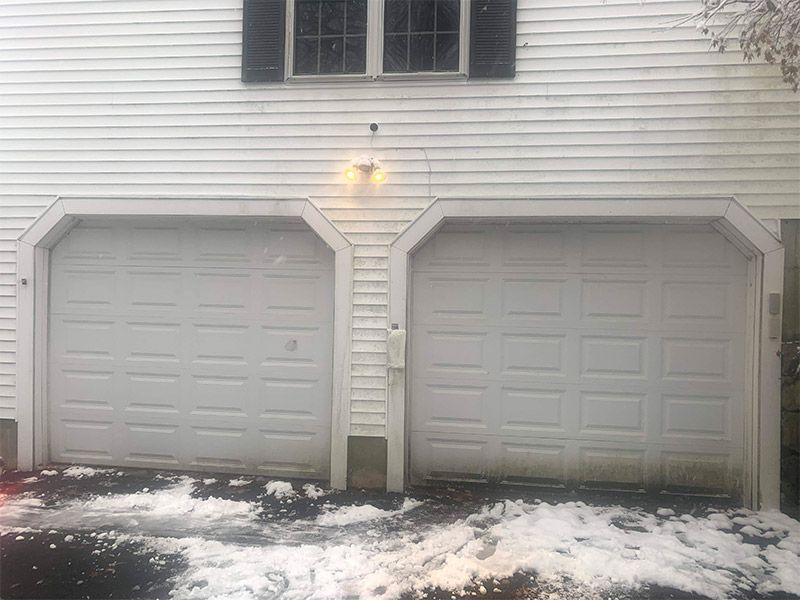 Two white garage doors on a house, with snow on the ground and a lit porch light.