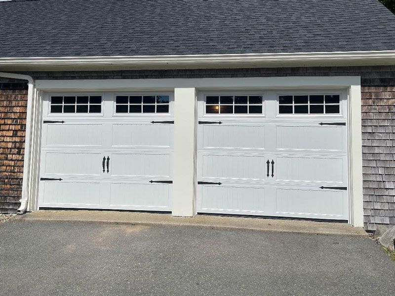 Two white garage doors with small windows, set in a shingled house exterior.