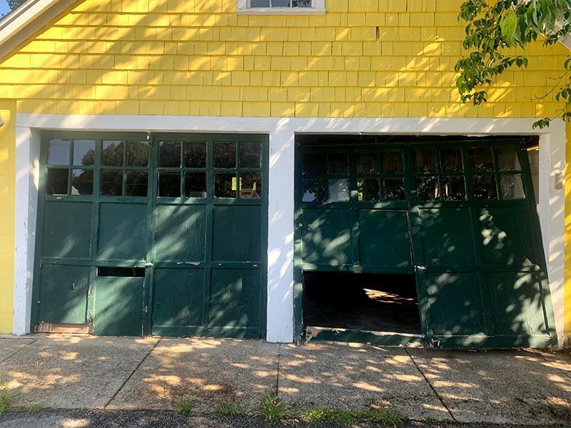 Yellow house front with two green garage doors, one partly open, beneath white trim and a leafy tree.
