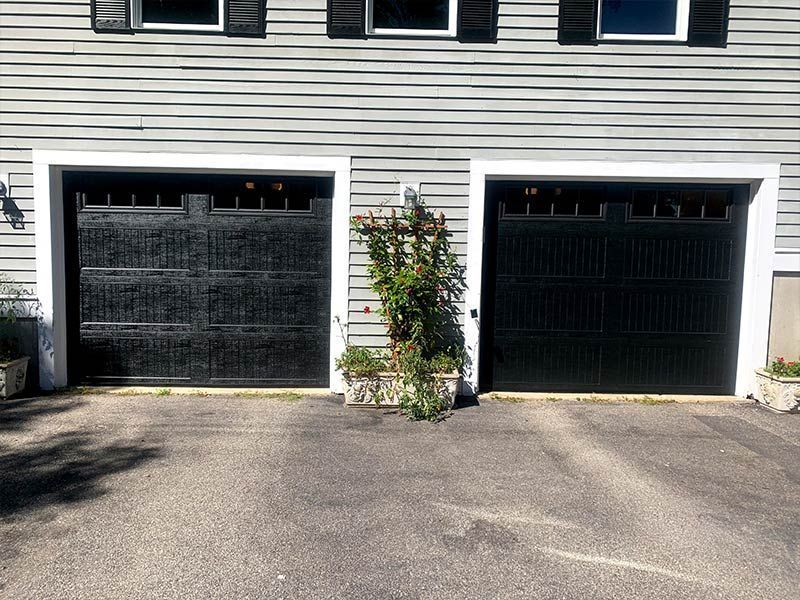Two black garage doors on a gray house with a small tree between them and a driveway in front