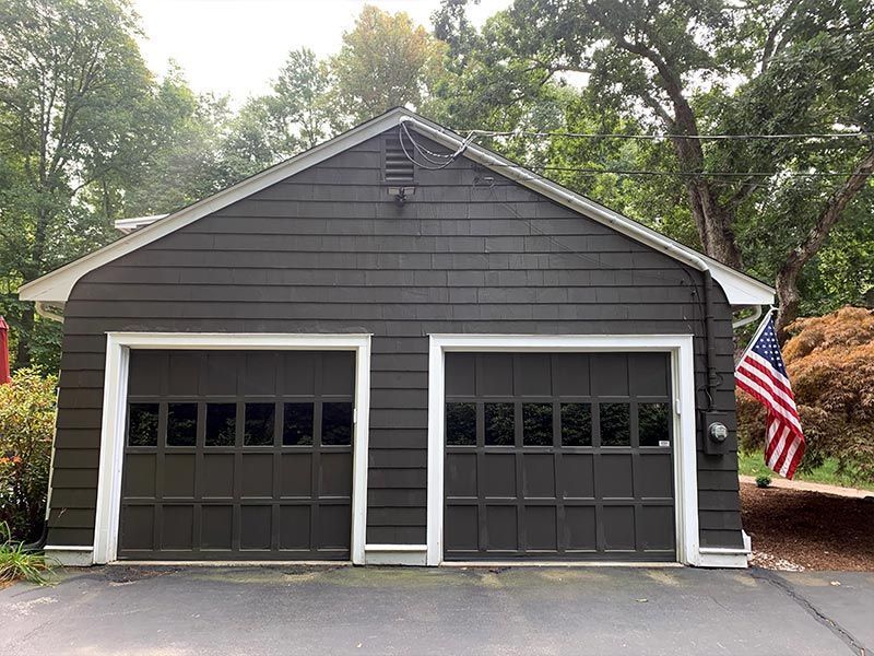 Gray two-car garage with white trim and an American flag in a wooded driveway