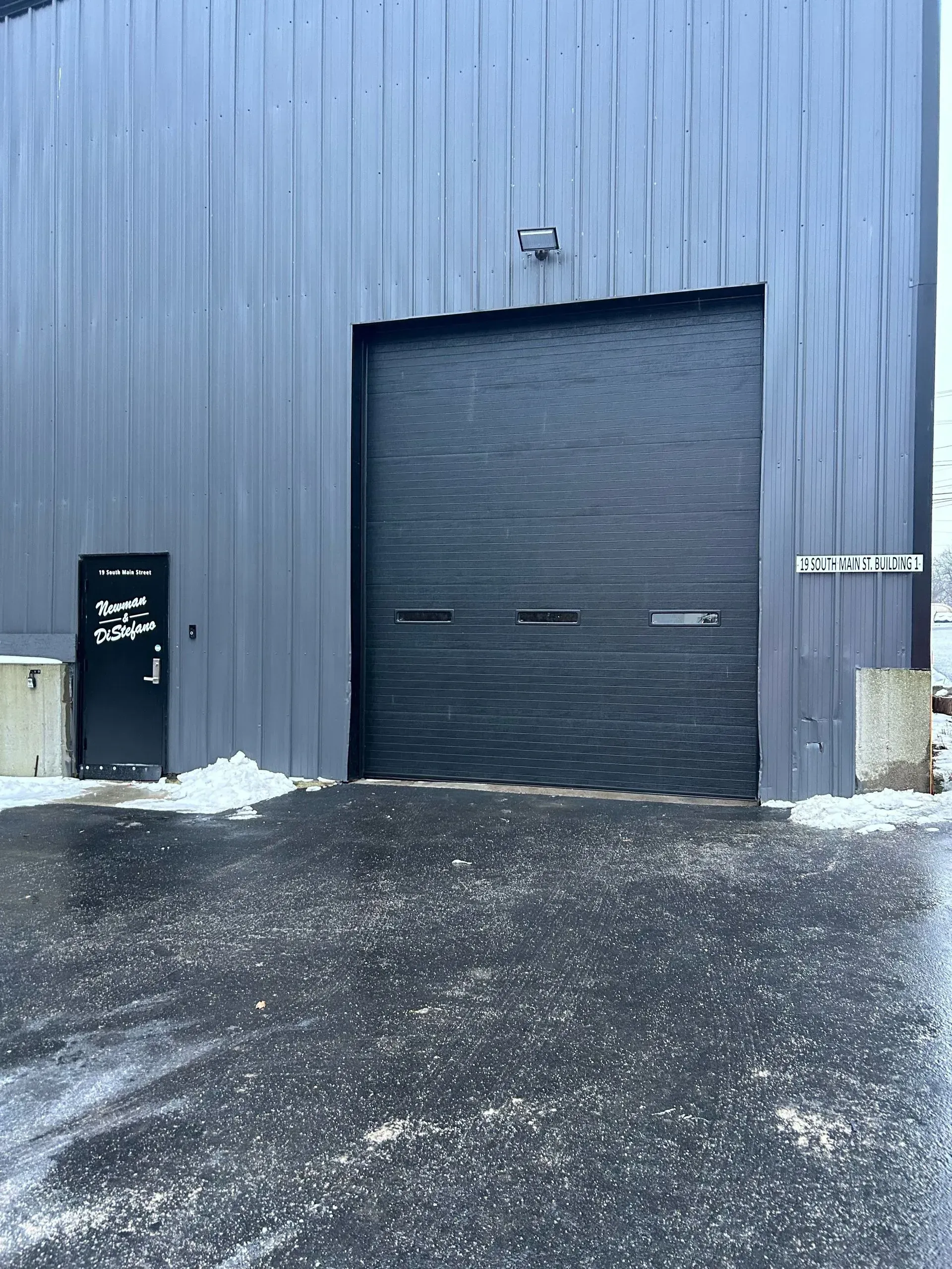 Blue industrial building with a large black garage door and snowy ground in front.