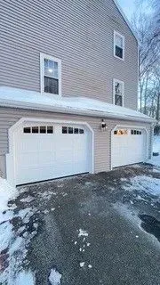 Snow-dusted driveway in front of a beige house with two white garage doors.