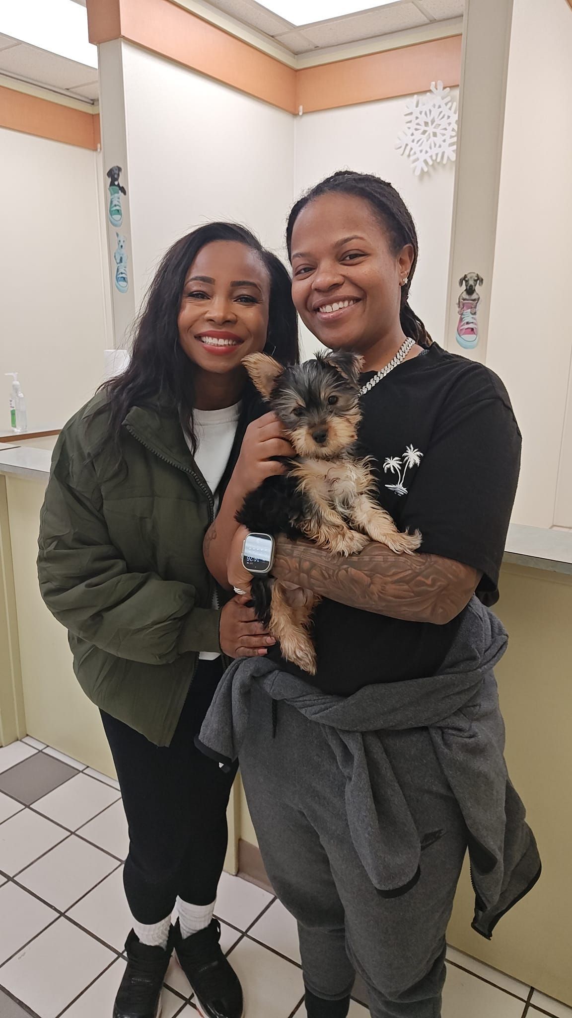 Two women smiling and holding a small dog in a hallway with beige walls and tiled floor.