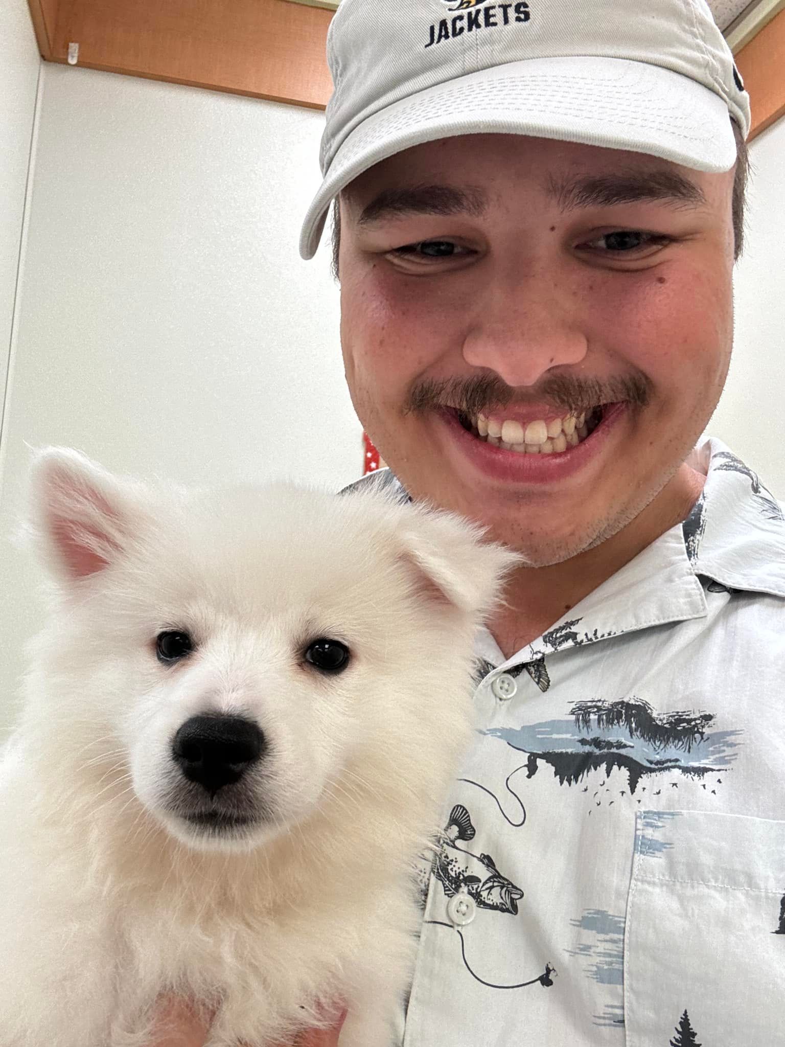 Smiling man in a white cap holding a fluffy white dog indoors
