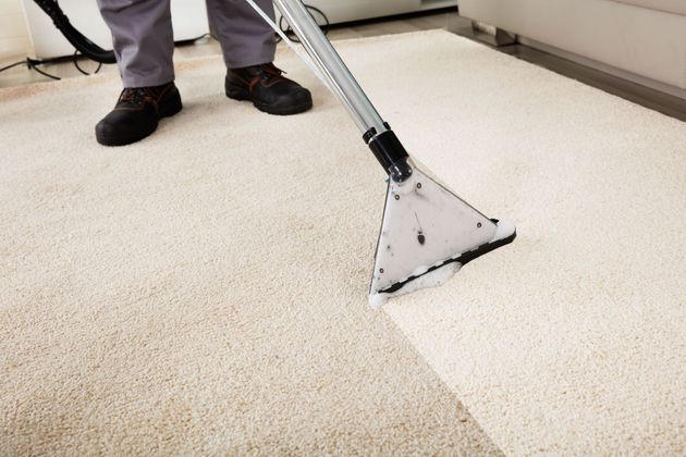Carpet cleaner vacuuming a beige carpet in a room, with a person’s legs visible.