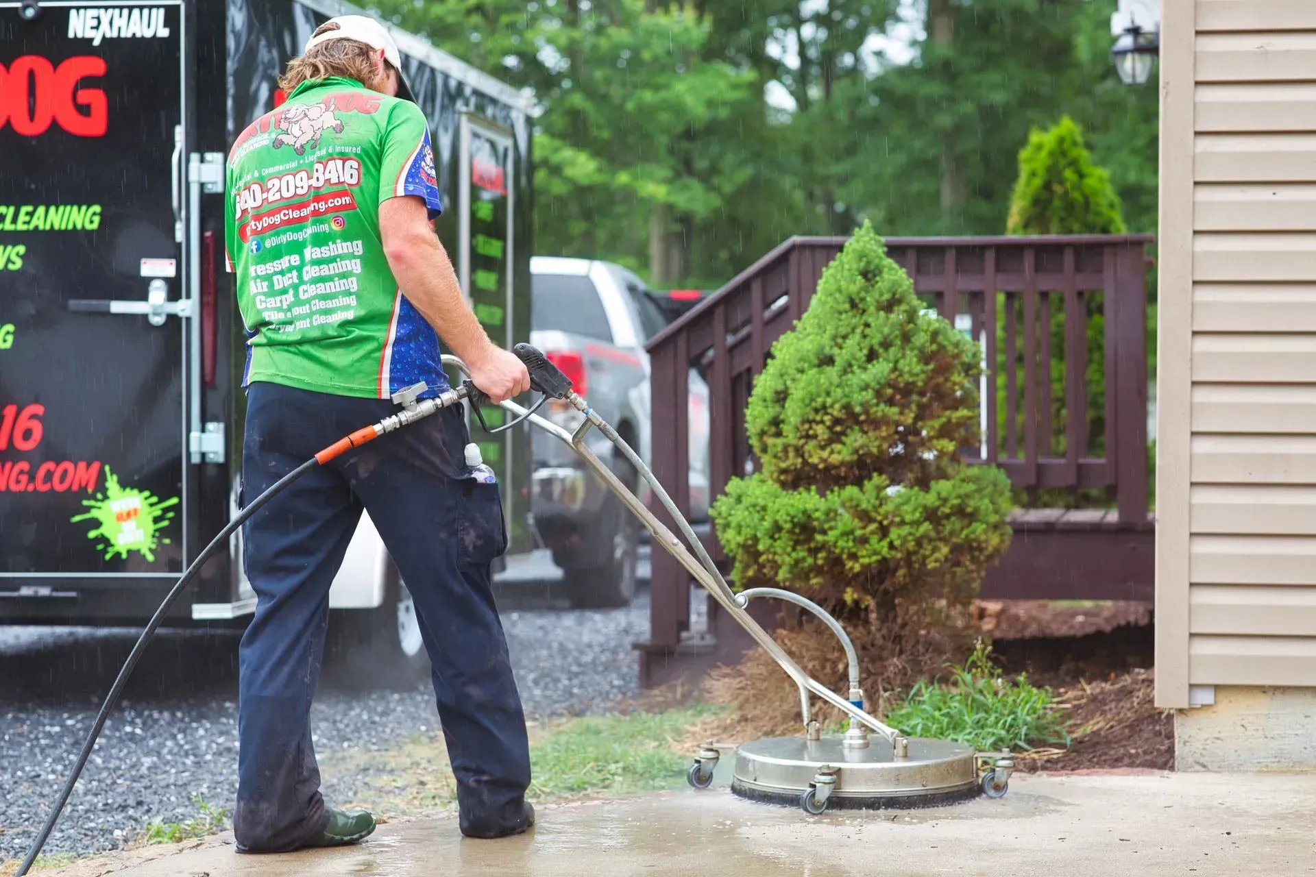 Worker using a pressure washer to clean a driveway beside a truck and shrubs
