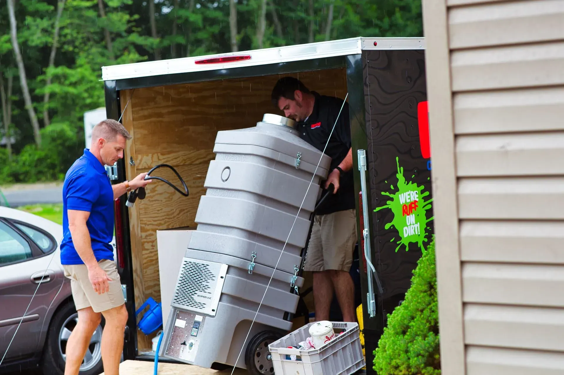 Two men moving a large gray cylinder from a shed beside a house