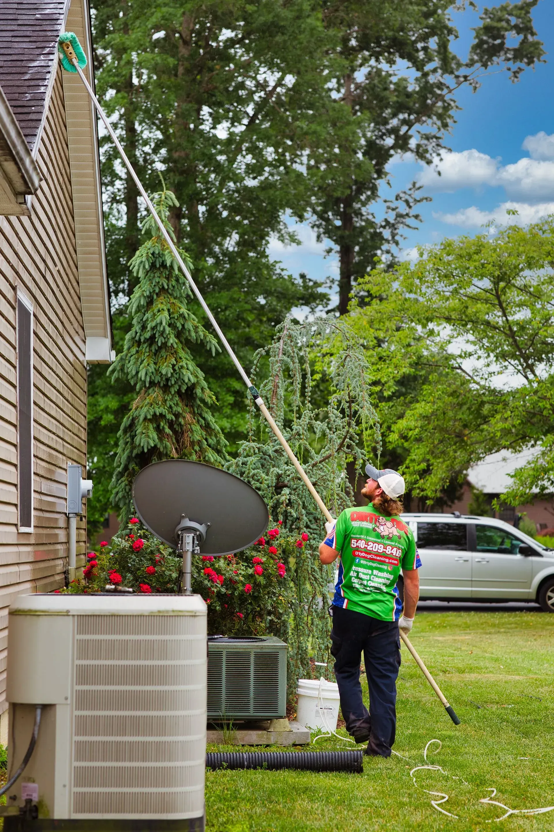 Worker trimming tree beside a brick house, with a satellite dish and parked car in the yard