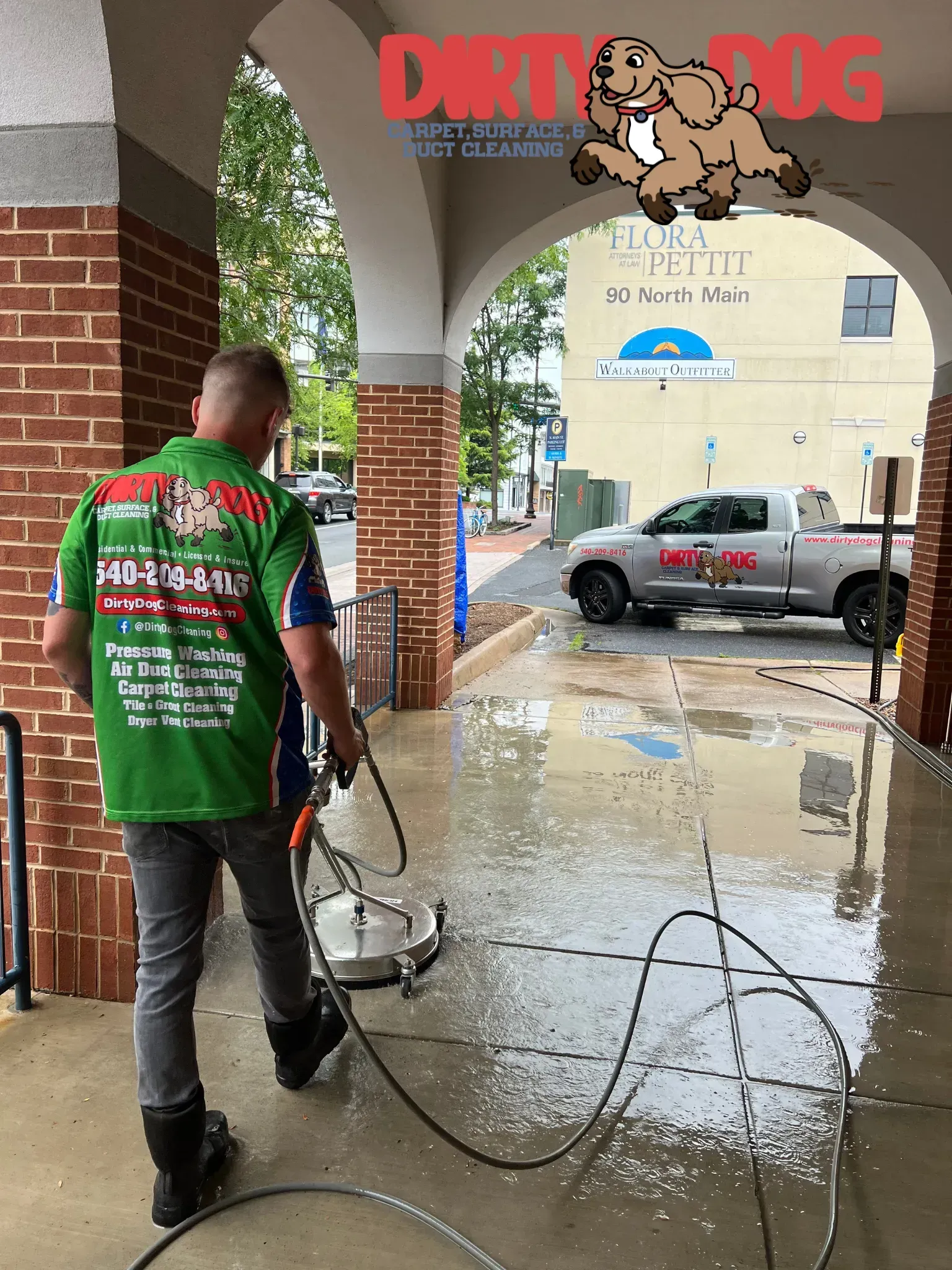 Worker pressure-washing a wet sidewalk under a brick arcade with a car parked outside during rain