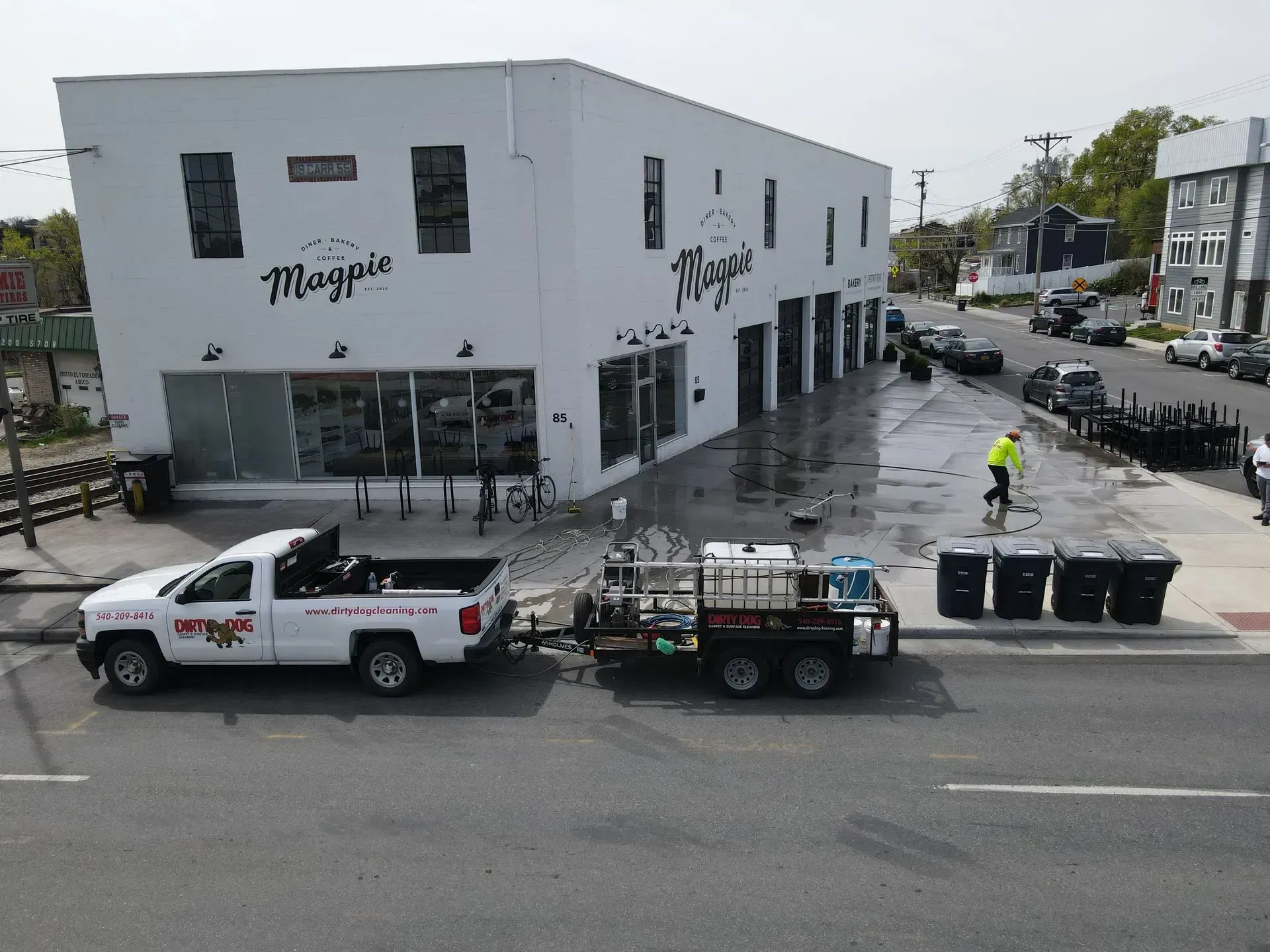 White corner building with trucks parked outside on a wet street, and a person in a yellow vest nearby