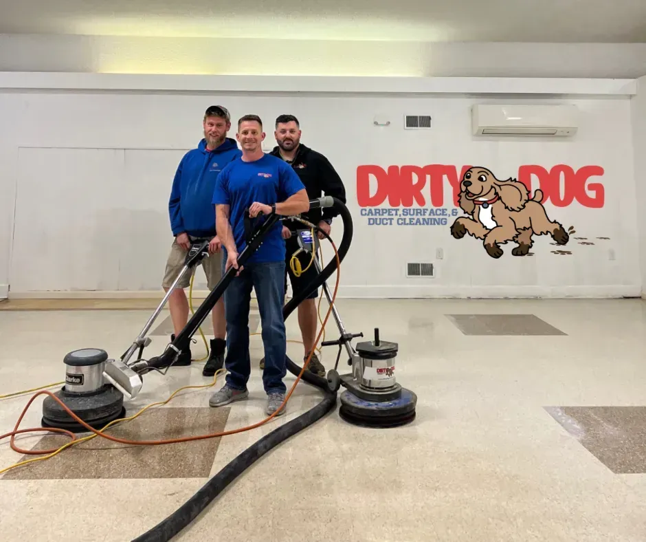 Three carpet cleaning workers in blue uniforms using floor machines in a room with a Dirty Dog Cleaners wall logo