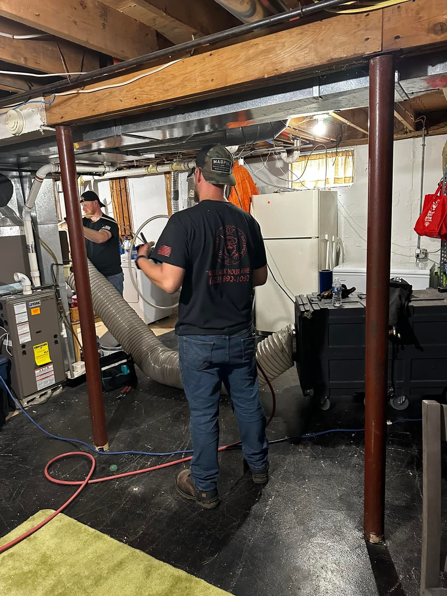 Two men working in a basement, near wooden beams and support poles. One holds a tool, the other works on a wall.