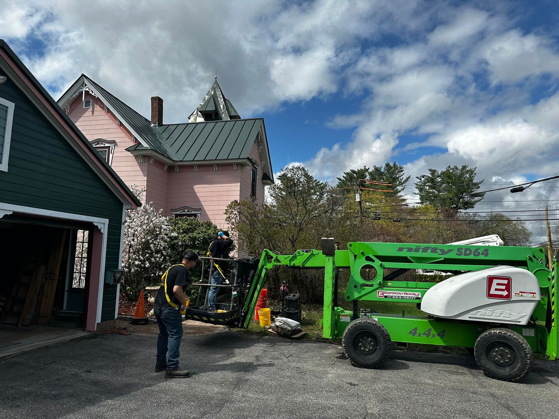 Construction workers near a pink house and green garage, using a green lift under a partly cloudy sky.